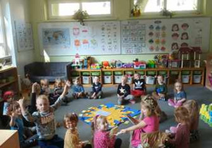 Zdjęcie przedstawia dzieci siedzące kręgu na dywanie podczas uroczystości urodzin, na środku widać pomoc edukacyjną "Słoneczko"