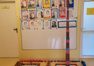 Zdjęcie przedstawia wystawę prac dzieci, na której znajdują się portrety Marii Montessori namalowane przez dzieci, różowa wieża, brązowe schody, czerwono niebieskie sztangi, czerwone sztangi, kubeczki z zafarbowaną na różne kolory wodą, tabliczki barwne, kolorowe cylindry, cylindry do osadzania, dwa napisy Maria Montessori ułożone z ruchomego alfabetu