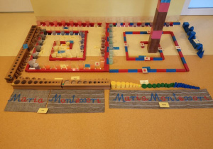 Zdjęcie przedstawia podłogę, na której znajdują się różowa wieża, brązowe schody, czerwono niebieskie sztangi, czerwone sztangi, kubeczki z zafarbowaną na różne kolory wodą, tabliczki barwne, kolorowe cylindry, cylindry do osadzania, dwa napisy Maria Montessori ułożone z ruchomego alfabetu