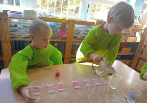 Dwóch chłopców siedzi przy stoliku. Przed nimi ułożone są plastikowe kubeczki i tabliczki barwne. Jeden z chłopców nalewa wodę z dzbanka do kubeczka