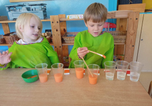 Chłopiec i dziewczynka siedzą przy stoliku. Chłopiec farbuje wodę nalaną w kubeczki na kolor pomarańczowy
