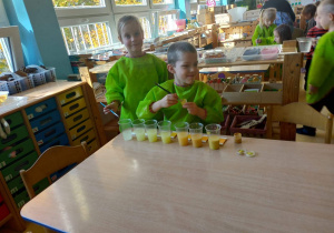 Chłopiec siedzi przy stoliku. Obok niego stoi dziewczynka. Chłopiec farbuje wodę nalaną w kubeczki na kolor żółty.