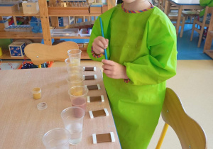 Dziewczynka siedzi przy stoliku i farbuje wodę nalaną w kubeczki na kolor brązowy