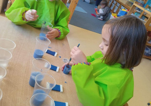 Dwie dziewczynki siedzą przy stoliku i farbują wodę nalaną w kubeczki na kolor niebieski. Za nimi dzieci bawiące się na dywanie.