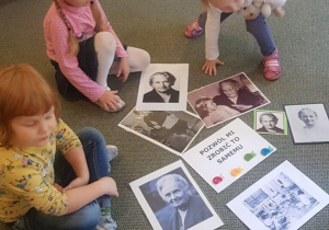Trzy dziewczynki siedzą na dywanie i przyglądają się portretom Marii Montessori oraz napisowi Pozwól mi zrobić to samemu