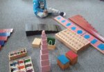 Chłopiec siedzi na dywanie. Przed nim znajduje się materiał rozwojowy taki jak czerwone sztangi, metalowe ramki, drewniane cylindry, puszki szmerowe, brązowe schody, różowa wieża, tabliczki barwne, cyfry szorstkie i czerwono niebieskie sztangi