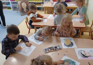 Dzieci siedzą przy stolikach i rysują portrety Marii Montessori
