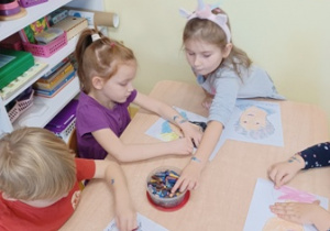 Dzieci siedzą przy stoliku i rysują portrety Marii Montessori, jedna z dziewczynek sięga po kredkę