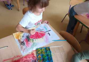 Chłopiec maluje tło swojej pracy plastycznej farbami