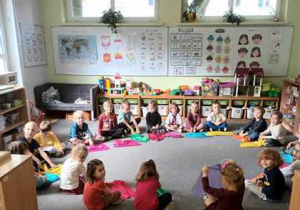 Dzieci siedzą na dywanie i wykonują improwizacje ruchowe kolorowymi chustkami do muzyki