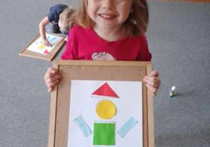 Dziewczynka trzyma w dłoniach podkładkę z pracą plastyczną przedstawiająca pajacyka ułożonego z figur geometrycznych