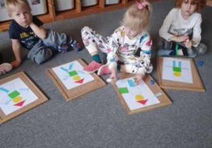 Dzieci siedzą na dywanie i układają na podkładkach kompozycję z figur geometrycznych