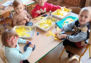 Dzieci siedzą przy stoliku i szykują się do przyklejania żółtych płatków słonecznika