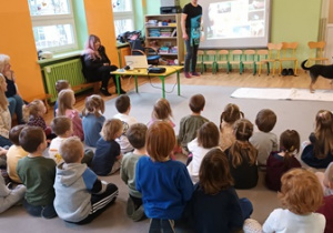 Dzieci siedzą na dywanie w sali gimnastycznej i słuchają prezentacji