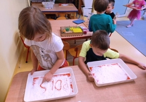 Dzieci ćwiczą pisanie na tacy wypełnionej mąką