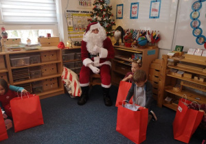 Mikołaj siedzi na krześle a dzieci oglądają prezenty, które dostały