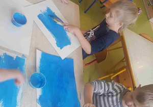 Dzieci malują niebieskie tło szerokim pędzlem na dużej płaszczyźnie