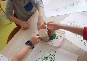 Dzieci tworzą choinkę z wykorzystaniem zielonych kół odbitych z papierowego kubeczka