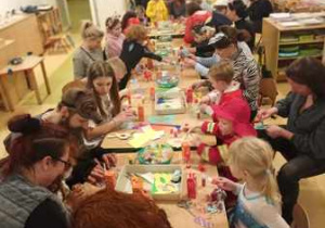 Dzieci wraz z rodzicami siedzą przy dużym stole i ozdabiają maski karnawałowe
