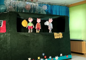 Zdjęcie przedstawia zieloną scenę, na której stoją kukiełki przedstawiające trzy świnki.