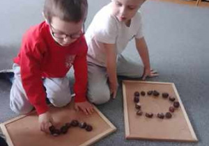 Chłopcy siedzą na dywanie i układają na podkładkach cyfry z kasztanów
