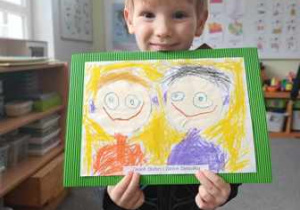 Chłopiec trzyma w dłoniach namalowany przez siebie portret Babci i Dziadka