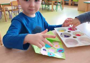 Chłopiec siedzi przy stoliku i stempluje kwiaty palcem umoczonym w farbie