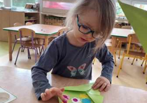 Dziewczynka siedzi przy stoliku i stempluje kwiaty palcami umoczonymi w farbie na swojej pracy plastycznej