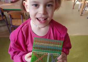 Dziewczynka pozuje do zdjęcia z wykonaną przez siebie pomocą edukacyjną - nawlekanie gumek według wzoru na geoplan