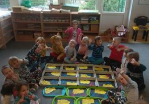 Dzieci siedzą na dywanie ze swoimi pracami plastycznymi przedstawiającymi samodzielnie sformułowane Prawa Dziecka.