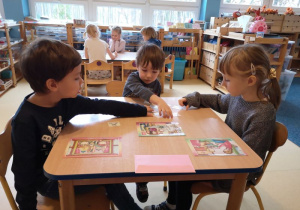 Dwóch chłopców i dziewczynka siedzą przy stoliku i dopasowują obrazki do ilustracji. W tle przy innym stoliku siedzą inne dzieci.
