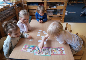 Trzy dziewczynki i jeden chłopiec siedzą przy stoliku i dopasowują obrazki do ilustracji. Jedna z dziewczynek wskazuje palcem postać taty.