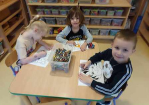 Dzieci starsze siedzą przy stoliku i kolorują pastelami sylwety zwierząt