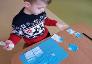 Chłopiec składa w całość zimową ilustrację i przykleja do niebieskiej kartki