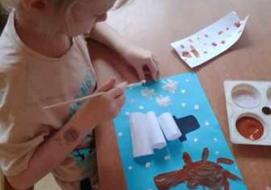 Dziewczynka trzyma w dłoniach pędzel umoczony w białej farbie i maluje śnieg na swojej pracy plastycznej