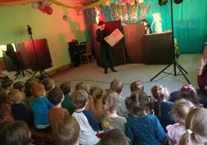 Na zdjęciu widać scenografię wystawianego przedstawienia, aktora oraz dzieci oglądające teatrzyk