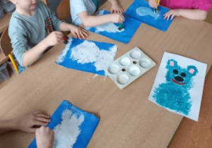 Dzieci siedzą przy stoliku i za pomocą gąbki umoczonej w białej farbie tworzą niedźwiadka polarnego