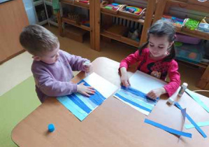 Dziewczynka i chłopiec przyklejają niebieskie paski kolorowego papieru na białej kartce