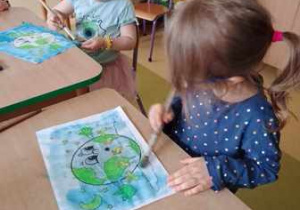 Dwie dziewczynki siedzą przy stoliku i malują niebieską akwarelą tło swojej pracy plastycznej