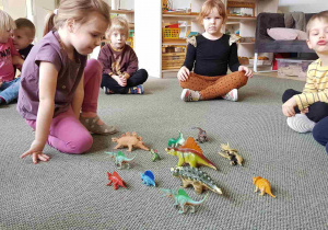 Dzieci siedzą na dywanie i rozpoznają figurki dinozaurów