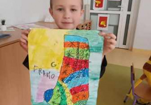 Chłopiec trzyma w dłoniach wykonaną przez siebie pracę plastyczną przedstawiającą kolorową skarpetkę