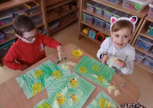 Dziewczynka i chłopiec doklejają żółte piórka do swojej pracy plastycznej