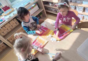 Dzieci przy stoliku w trakcie wykonywania pracy plastycznej doklejają paski szarego papieru
