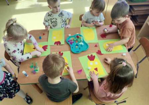 Dzieci siedzą przy stoliku i doklejają elementy do swojej pracy plastycznej prezentującej kurczaczki
