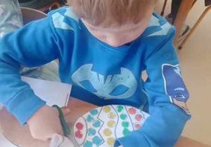 Chłopiec siedzi przy stoliku i wycina pisankę wielkanocną wyklejoną przez siebie plasteliną