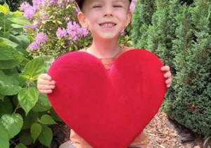 Tytus w ogrodzie przedszkolnym pozuje do zdjęcia trzymając w dłoniach serce dla Mamy i Taty