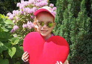 Antosia w ogrodzie przedszkolnym pozuje do zdjęcia trzymając w dłoniach serce dla Mamy i Taty