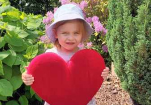 Dziewczynka w ogrodzie przedszkolnym pozuje do zdjęcia trzymając w dłoniach serce dla Mamy i Taty