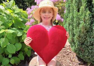 Anielka w ogrodzie przedszkolnym pozuje do zdjęcia trzymając w dłoniach serce dla Mamy i Taty