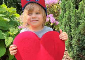 Jeremi w ogrodzie przedszkolnym pozuje do zdjęcia trzymając w dłoniach serce dla Mamy i Taty